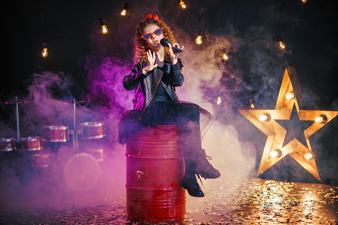 Beautiful girl with curly hair wearing leather jacket, red sunglasses sing into a wireless microphone for karaoke while sitting on red tank in recording studio or stage cours chant enfants